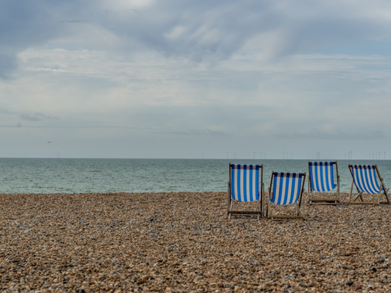 Four blue-and-white striped beach chairs on a pebbled shore facing the calm sea under a cloudy sky nearby.