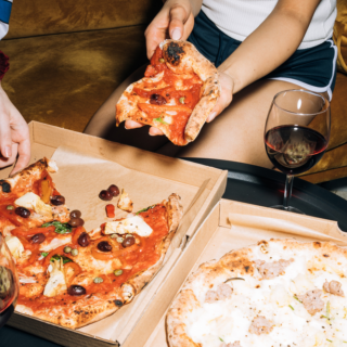 People sit on a sofa sharing two pizzas, with glasses of red wine on a coffee table and pizza slices being handed around.