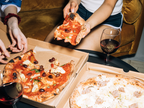 People sit on a sofa sharing two pizzas, with glasses of red wine on a coffee table and pizza slices being handed around.