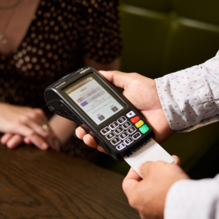 Person using a handheld card payment terminal to process a sale, swiping a card while a customer sits nearby at a wooden counter.
