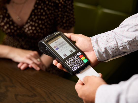 Person using a handheld card payment terminal to process a sale, swiping a card while a customer sits nearby at a wooden counter.