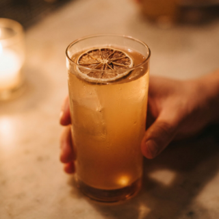 Tall glass of iced beverage with a lemon wheel on top, held by two hands at a marble bar counter with warm candlelight in the background