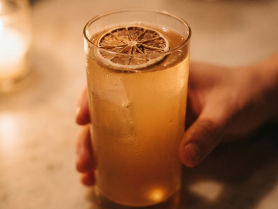 Tall glass of iced beverage with a lemon wheel on top, held by two hands at a marble bar counter with warm candlelight in the background