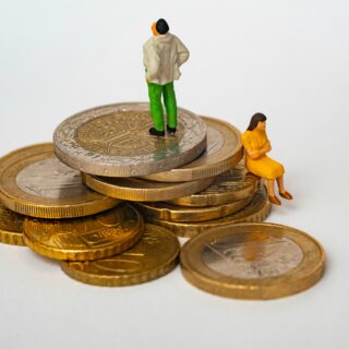 Tiny figurines atop a stack of coins: a man in green pants and a beige jacket stands while a small orange-dressed woman sits at the edge of the coins.