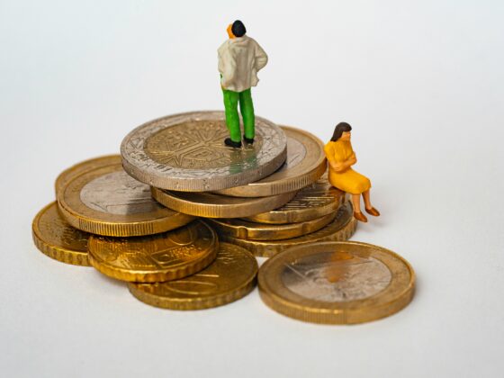 Tiny figurines atop a stack of coins: a man in green pants and a beige jacket stands while a small orange-dressed woman sits at the edge of the coins.