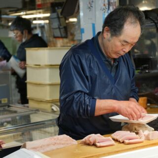 Man in a blue jacket slicing fish on a wooden board at a market counter, with sashimi pieces laid out nearby in a seafood shop setting.