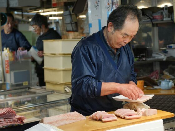 Man in a blue jacket slicing fish on a wooden board at a market counter, with sashimi pieces laid out nearby in a seafood shop setting.