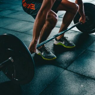 Close-up of a person performing a deadlift, gripping a loaded barbell on a rubber gym floor, wearing gray Nike shoes with neon green soles.