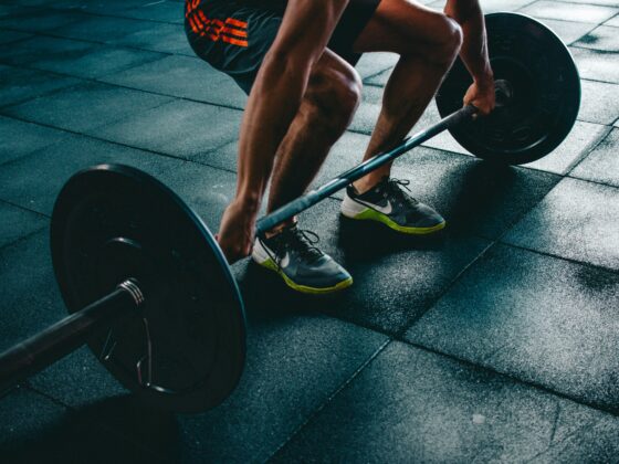 Close-up of a person performing a deadlift, gripping a loaded barbell on a rubber gym floor, wearing gray Nike shoes with neon green soles.