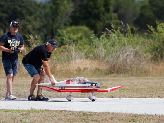 Two men with a remote-controlled model airplane on a sunny field; one holds a controller while the other adjusts the plane's nose.