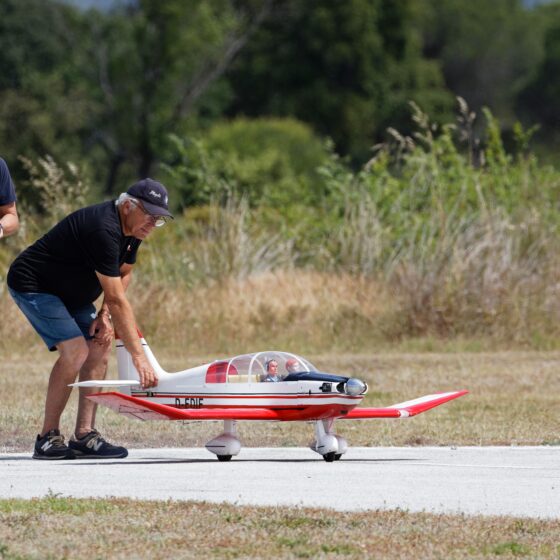 Two men with a remote-controlled model airplane on a sunny field; one holds a controller while the other adjusts the plane's nose.