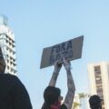 Person with tattooed arms raises a cardboard sign reading 'FORA BOZO' at a street protest, with a crowd and tall buildings in the background.