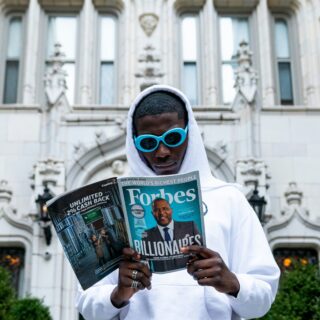 Person wearing a white hoodie and bright blue sunglasses reads a Forbes magazine outdoors in front of a historic stone building with ornate arches.