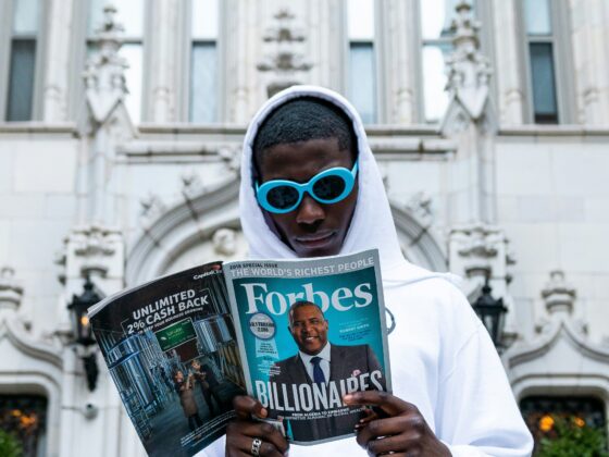 Person wearing a white hoodie and bright blue sunglasses reads a Forbes magazine outdoors in front of a historic stone building with ornate arches.