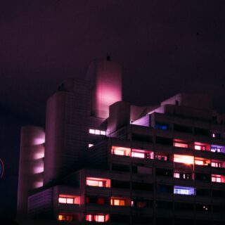 Night shot of a modern building with pink and purple illuminated windows against a dark sky.