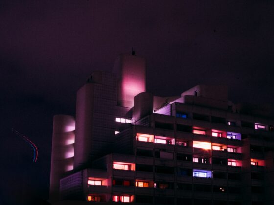 Night shot of a modern building with pink and purple illuminated windows against a dark sky.