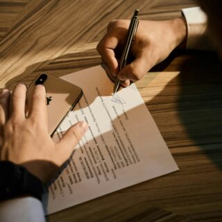 Close-up of a hand signing a document with a pen on a wooden desk, with a smartphone nearby.
