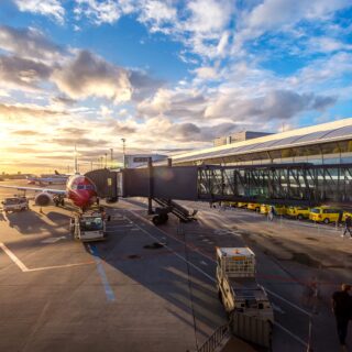 Sunset at an airport gate with a red airplane docked to a jet bridge and ground crews nearby