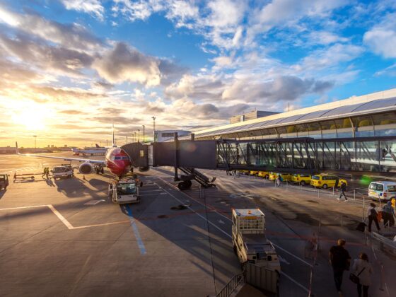 Sunset at an airport gate with a red airplane docked to a jet bridge and ground crews nearby