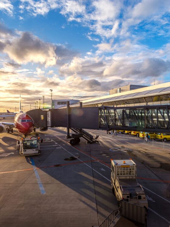 Sunset at an airport gate with a red airplane docked to a jet bridge and ground crews nearby