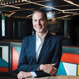 Man in a navy suit and white shirt leaning on a wooden counter, smiling in a colorful, modern venue.