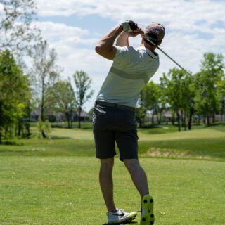 Male golfer in a light gray shirt and cap mid-swing on a sunny golf course, trees in the background.