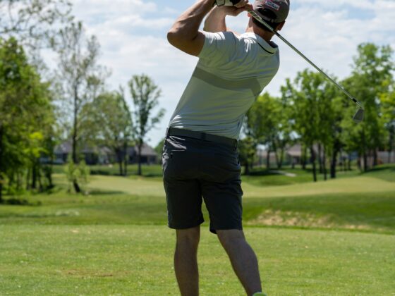 Male golfer in a light gray shirt and cap mid-swing on a sunny golf course, trees in the background.