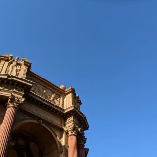 Stone classical building corner with carved frieze, ornate pediment and tall red columns under a clear blue sky, statues atop the cornice.