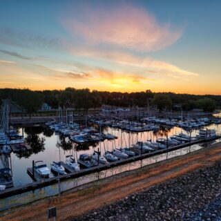 Sunset over a harbor with moored boats, calm water, and a tree-lined shore in the background.