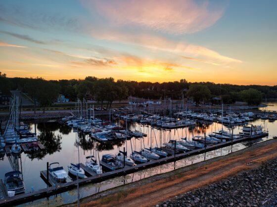 Sunset over a harbor with moored boats, calm water, and a tree-lined shore in the background.
