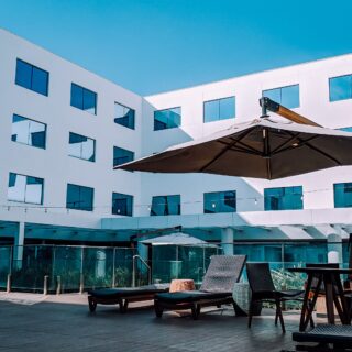 Outdoor seating area with wicker lounge chairs and a large umbrella in front of a white modern building with blue-tinted windows.