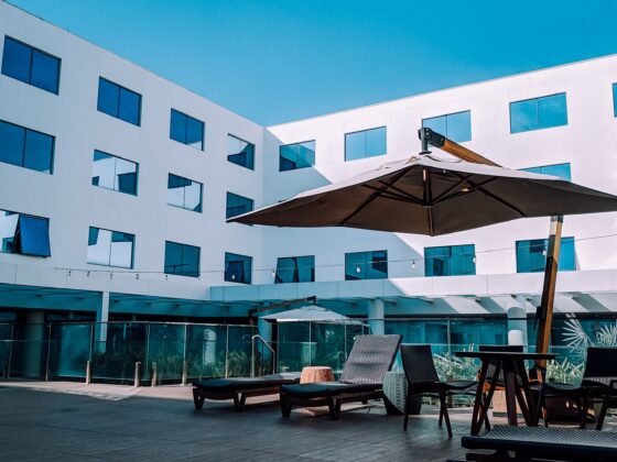 Outdoor seating area with wicker lounge chairs and a large umbrella in front of a white modern building with blue-tinted windows.