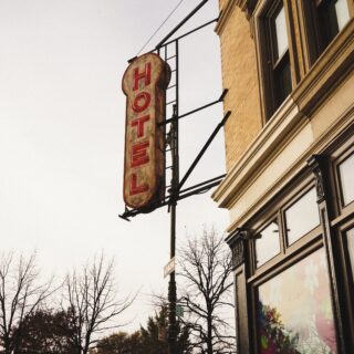 Vertical red 'HOTEL' sign mounted on a metal frame beside a brick storefront on a city street, with bare trees in the background and a stop/one-way sign nearby.