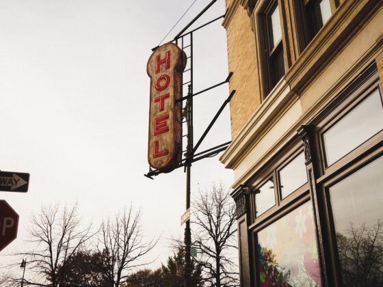 Vertical red 'HOTEL' sign mounted on a metal frame beside a brick storefront on a city street, with bare trees in the background and a stop/one-way sign nearby.
