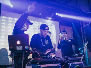 Two DJs behind a mixer and laptops on a club table, hands adjusting controls under blue stage lights.