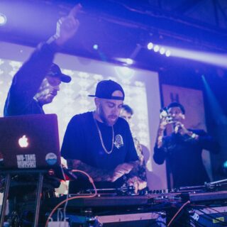 Two DJs behind a mixer and laptops on a club table, hands adjusting controls under blue stage lights.