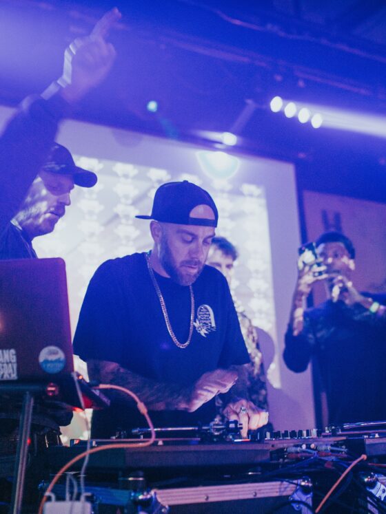 Two DJs behind a mixer and laptops on a club table, hands adjusting controls under blue stage lights.