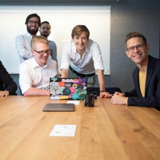 A group of coworkers gathered around a wooden conference table, smiling at the camera in a modern office.