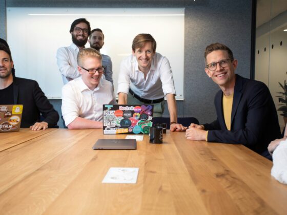 A group of coworkers gathered around a wooden conference table, smiling at the camera in a modern office.