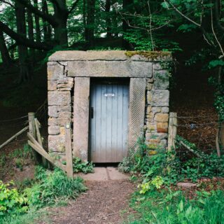 Stone-walled entrance in a wooded hillside with a gray metal door and moss on top, surrounded by a barbed-wire fence.