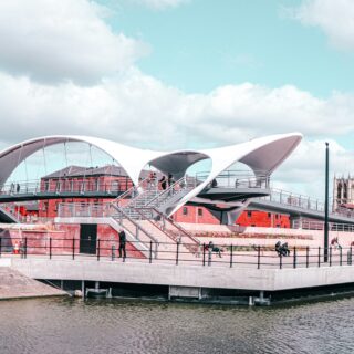 Modern white curved pedestrian bridge over a river with brick buildings and a church tower in the background.