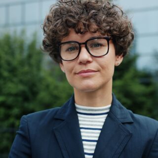 Person with short curly hair wearing large black glasses, a striped shirt, and a navy blazer, posing outdoors in front of greenery and a building.