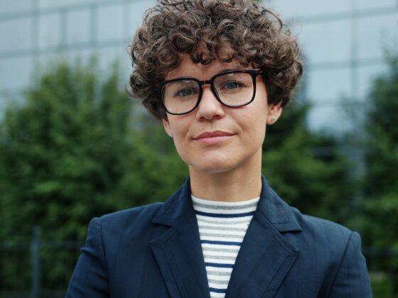 Person with short curly hair wearing large black glasses, a striped shirt, and a navy blazer, posing outdoors in front of greenery and a building.