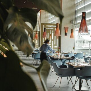 Restaurant dining area with blue chairs and wooden tables; a server arranges flowers while warm orange pendant lights hang above, and large windows overlook a marina with boats.
