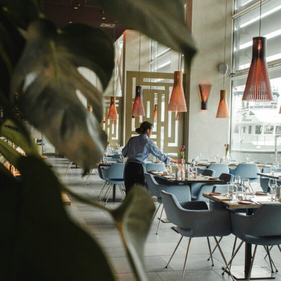 Restaurant dining area with blue chairs and wooden tables; a server arranges flowers while warm orange pendant lights hang above, and large windows overlook a marina with boats.