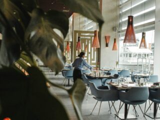 Restaurant dining area with blue chairs and tables set with glassware; a server in a blue uniform tends a table near large windows and orange pendant lamps.
