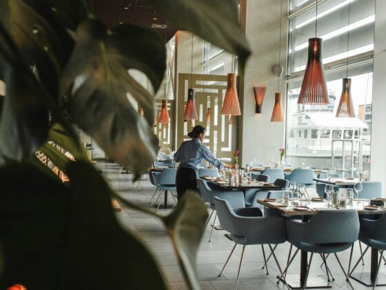 Restaurant dining area with blue chairs and tables set with glassware; a server in a blue uniform tends a table near large windows and orange pendant lamps.