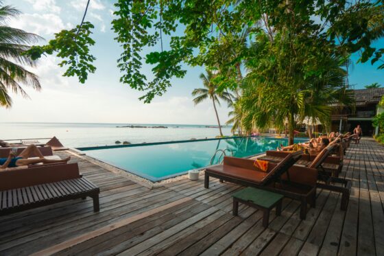 Poolside lounge area on a wooden deck with palm trees, overlooking calm sea and distant rocks at a tropical beach resort