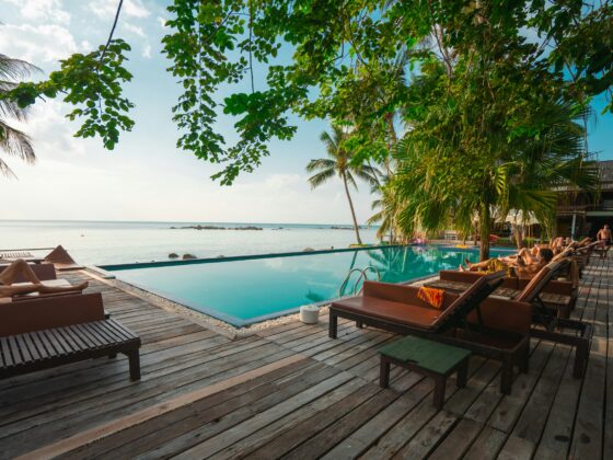 Poolside lounge area on a wooden deck with palm trees, overlooking calm sea and distant rocks at a tropical beach resort