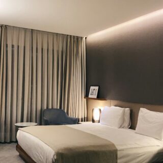 Hotel bedroom with a low wooden bed frame, white bedding, and a beige runner; a night lamp glows beside a framed photo on a shelf.